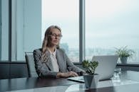 A confident businesswoman in a grey suit working on a laptop in a modern office setting.